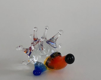 May include: A glass hedgehog figurine with a clear body, orange, blue, and yellow accents, and a black nose.