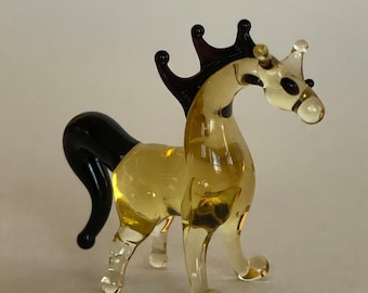May include: A small glass horse figurine with a yellow body, black tail and mane, and a purple crown on its head.
