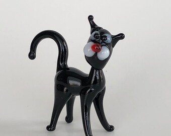 May include: A small black glass cat figurine featuring a long curved tail, two white circular cheek accents, a small red nose, and two light blue eyes. The cat stands on four slender legs with its head turned slightly to the right side.