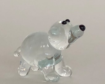 May include: A clear glass figurine of a dog with black eyes and nose. The dog is in a standing position, with its head tilted upwards. The figurine is small and appears to be handmade.