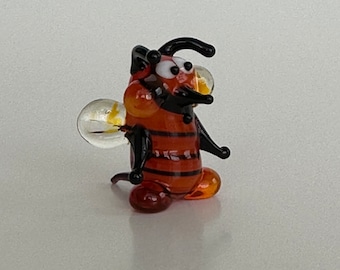 May include: A small glass figurine of a cartoon bee with black and white stripes, brown and orange body, and black eyes. The bee has a yellow and clear glass wing.