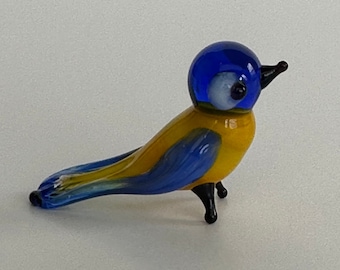 May include: A small glass bird figurine with a blue head, yellow body, and blue wings. The bird has black legs and a white eye.