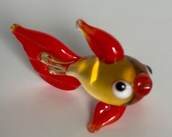 May include: A small glass figurine of a fish with a translucent yellow body and vibrant red fins. The fish features a white eye with a black pupil, a rounded red mouth, and a long, flowing red tail. The glass material is smooth and glossy.