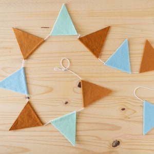 May include: A string of felt triangle flags in alternating colours of light blue, mint green, and orange brown. The flags are strung together with white string.