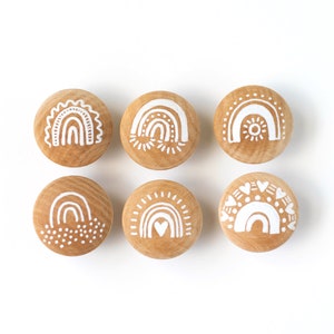 Peut inclure: Six boutons en bois avec différents motifs d'arc-en-ciel peints en blanc. Les motifs comprennent un arc-en-ciel avec un soleil, un arc-en-ciel avec des points, un arc-en-ciel avec un cœur et un arc-en-ciel avec des cœurs.