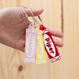 May include: Three crayon-shaped keychains in pink, yellow, and red, each with a name printed on them: Madeline, Bella, and Piper. The keychains are attached to silver keyrings.