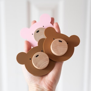 Puede incluir: Tres ganchos de pared o pomos de cajón con forma de oso en rosa claro y dos tonos de marrón. Cada pieza presenta una base plana con forma de oso y una pieza de madera clara circular en el centro que representa el hocico, con detalles negros.