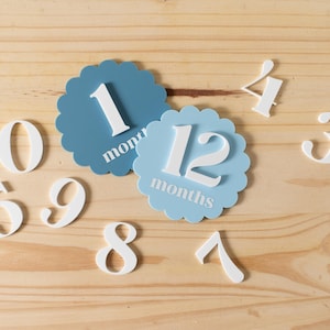 K&ouml;nnte beinhalten: Blaue und wei&szlig;e Baby-Meilenstein-Karten mit den Zahlen 1, 12, 3, 4, 5, 6, 7, 8, 9 und 0. Die Karten liegen auf einem hellen Holzhintergrund.