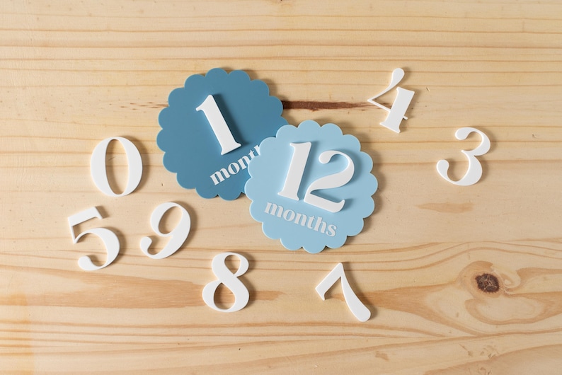 K&ouml;nnte beinhalten: Blaue und wei&szlig;e Baby-Meilenstein-Karten mit den Zahlen 1, 12, 3, 4, 5, 6, 7, 8, 9 und 0. Die Karten liegen auf einem hellen Holzhintergrund.