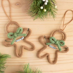 May include: Two handmade gingerbread man Christmas ornaments. Each ornament is crocheted in a tan yarn, with a green bow and a name spelled out in white beads. The ornaments are hanging from a tan string.