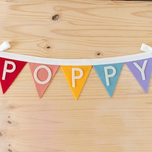 May include: A felt banner with the word "POPPY" spelled out in white letters on colorful triangular flags. The flags are red, pink, yellow, blue, and purple.