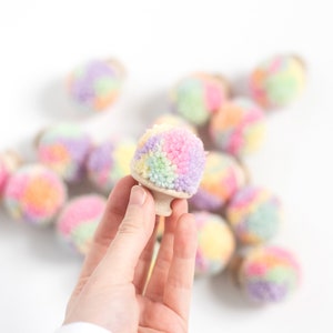 Könnte beinhalten: Eine Hand hält einen pastellfarbenen Regenbogen-Pompon-Anhänger. Der Anhänger ist aus Garn gefertigt und hat eine Holzbasis. Im Hintergrund befinden sich weitere pastellfarbene Regenbogen-Pompon-Anhänger.