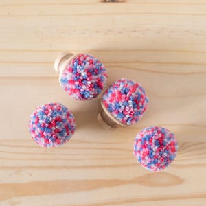 May include: Four colorful pom-poms with a pink, blue, and white color scheme. The pom-poms are attached to wooden bases.