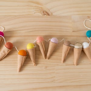 Op de afbeelding: Een slinger met negen vilten ijshoorntjes in diverse kleuren, waaronder roze, oranje, geel, perzik, lavendel, bruin, beige, wit en groenblauw, die allemaal aan een dun wit koord zijn geregen voor decoratieve ophangdoeleinden.