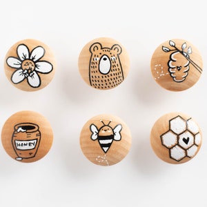 Może przedstawiać: Sześć drewnianych gałek z różnymi ilustracjami o tematyce pszczół. Gałki przedstawiają stokrotkę, niedźwiedzia, plaster miodu, pszczołę, słoik miodu i plaster miodu z sercem.