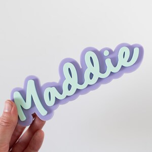 Może przedstawiać: Lawendowy wycięty napis z imieniem "Maddie" w jasnozielonej czcionce. Nazwa jest ułożona na tle w kształcie chmury. Wycięcie jest trzymane przez dłoń.