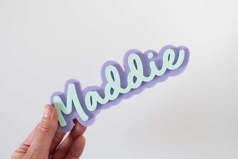 Może przedstawiać: Lawendowy wycięty napis z imieniem "Maddie" w jasnozielonej czcionce. Nazwa jest ułożona na tle w kształcie chmury. Wycięcie jest trzymane przez dłoń.