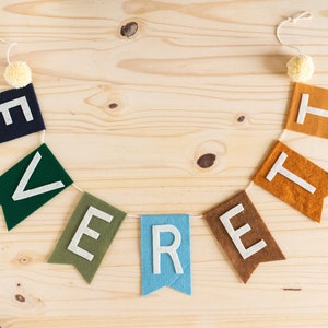 May include: A felt banner with the word "EVERETT" spelled out in white letters on colourful felt flags. The flags are strung together with white twine and have pom-poms on each end.