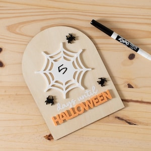 Puede incluir: Un letrero de cuenta regresiva de Halloween de madera con un diseño de telaraña blanca, el número 5 y las palabras "días para Halloween" en blanco y naranja. Incluye decoraciones de arañas negras y un marcador blanco y negro.