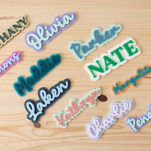 Może przedstawiać: Kolekcja kolorowych wycięć z imionami w r&oacute;żnych czcionkach i kolorach, w tym "Bethany", "Olivia", "Parker", "Nate", "Simmons", "Maddie", "Laken", "Kathryn", "Charlie", "Penelope" i "Margarita". Wycięcia ułożone są na jasnej drewnianej powierzchni.