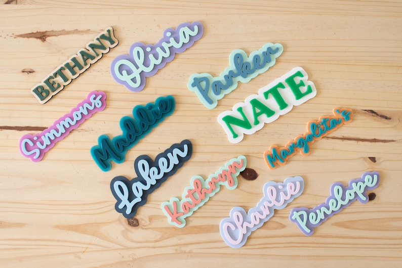 Może przedstawiać: Kolekcja kolorowych wycięć z imionami w r&oacute;żnych czcionkach i kolorach, w tym "Bethany", "Olivia", "Parker", "Nate", "Simmons", "Maddie", "Laken", "Kathryn", "Charlie", "Penelope" i "Margarita". Wycięcia ułożone są na jasnej drewnianej powierzchni.
