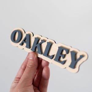 Może przedstawiać: Drewniany znak z napisem "OAKLEY" w ciemnoniebieskich literach. Litery są ułożone na jasnej drewnianej podstawie z falistą krawędzią. Znak jest trzymany na gładkim białym tle.