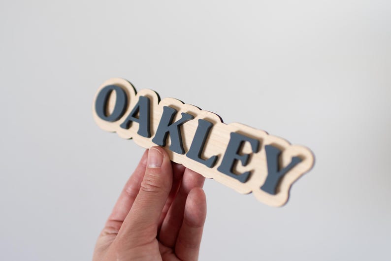 Może przedstawiać: Drewniany znak z napisem "OAKLEY" w ciemnoniebieskich literach. Litery są ułożone na jasnej drewnianej podstawie z falistą krawędzią. Znak jest trzymany na gładkim białym tle.