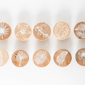 Puede incluir: Diez discos de madera redondos con diseños de hojas y flores tropicales pintados en blanco.