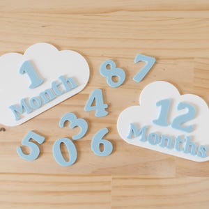 Pode incluir: Adereços fotográficos de marcos para bebês em madeira. Duas placas brancas em forma de nuvem com os números 1 e 12, e a palavra "Mês" em azul claro. Números adicionais em azul claro de 0 a 8 estão espalhados pelas nuvens.