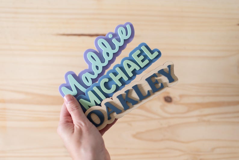 Może przedstawiać: Trzy spersonalizowane znaki z imionami. Imiona to "Maddie", "Michael" i "Oakley". "Maddie" jest fioletowy z jasnozielonym obrysem. "Michael" jest jasnozielony z niebieskim obrysem. "Oakley" jest granatowy z jasno brązowym obrysem.