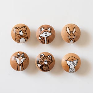 Puede incluir: Un juego de seis pomos redondos de madera, cada uno con una ilustración pintada a mano de un animal del bosque diferente. La colección incluye un oso, un zorro, un conejo, un ciervo, un mapache y un búho, en tonos marrón, blanco y negro.