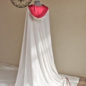 May include: A white hooded cloak with a red satin lining and trim. The cloak is draped over a mannequin, showcasing its flowing design. A decorative metal wall hanging is in the background.