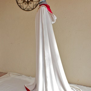 May include: A long, flowing white cloak with a red trim and a matching red collar. The cloak is draped over a mannequin, showcasing its elegant design. A decorative metal wall hanging is in the background.