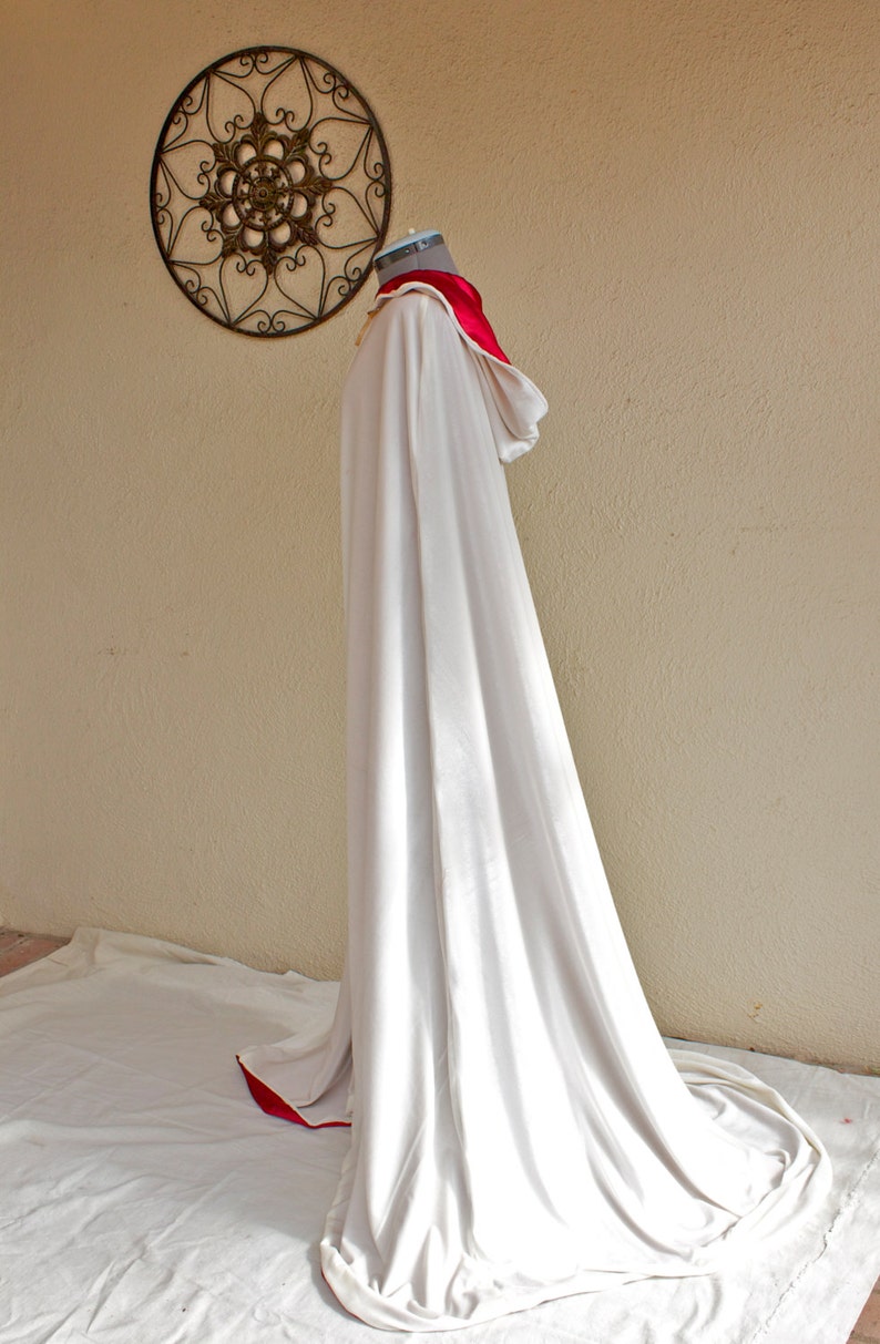 May include: A long, flowing white cloak with a red trim and a matching red collar. The cloak is draped over a mannequin, showcasing its elegant design. A decorative metal wall hanging is in the background.