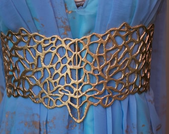 May include: A wide gold belt with an intricate, organic, web-like cutout pattern worn over a light blue dress featuring a subtle gold-toned mottled texture. The belt has a structured, rigid appearance and sits horizontally across the waist area.