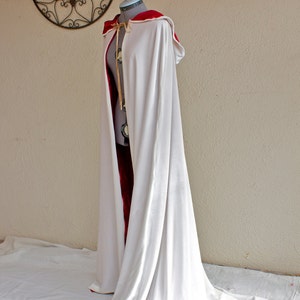 May include: A white cloak with a red-lined hood and a long train, displayed on a mannequin. The cloak is made of a flowing fabric and is secured with a gold-colored clasp. The background features a decorative wall hanging.