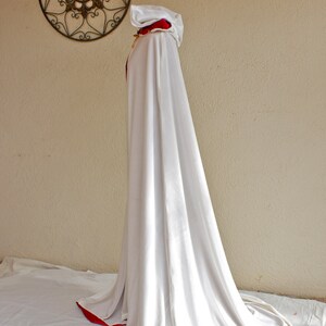 May include: A long, white hooded cloak with a red trim. The cloak is draped over an unseen form, creating a flowing silhouette. A decorative metal wall hanging is visible in the background.