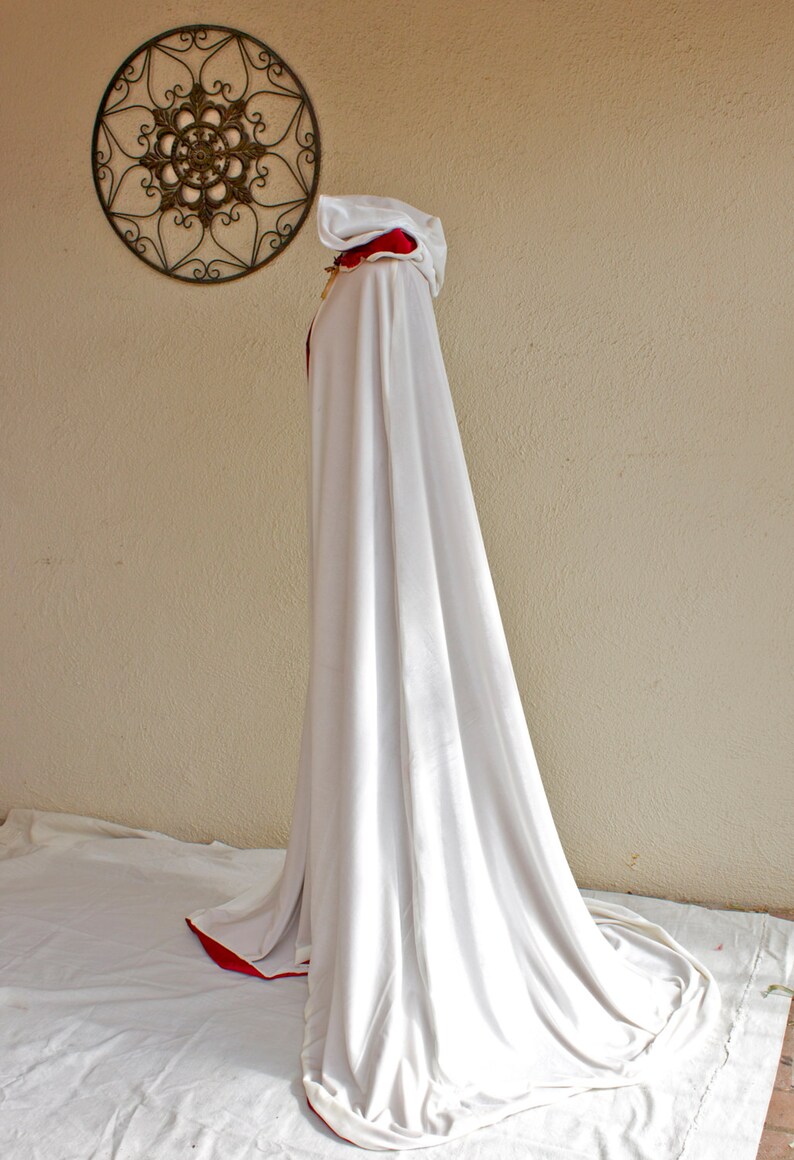May include: A long, white hooded cloak with a red trim. The cloak is draped over an unseen form, creating a flowing silhouette. A decorative metal wall hanging is visible in the background.