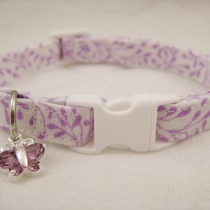 May include: A white and purple floral patterned cat collar with a white plastic buckle. A silver charm in the shape of a butterfly with purple gems hangs from the collar. The collar is approximately 12 inches long.