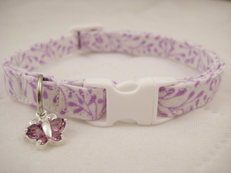 May include: A white and purple floral patterned cat collar with a white plastic buckle. A silver charm in the shape of a butterfly with purple gems hangs from the collar. The collar is approximately 12 inches long.