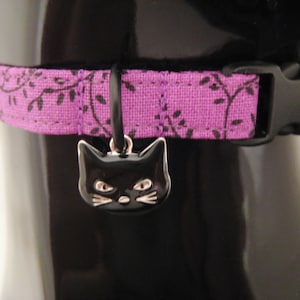 May include: A purple cat collar with a black plastic buckle and a black cat charm. The collar has a floral pattern in black. The cat charm is black with silver details.