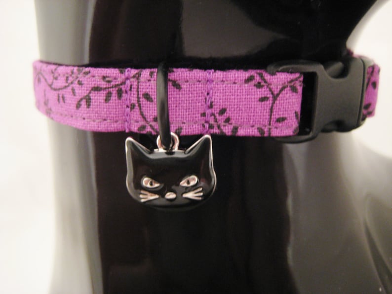 May include: A purple cat collar with a black plastic buckle and a black cat charm. The collar has a floral pattern in black. The cat charm is black with silver details.