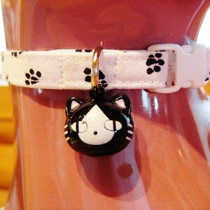 K&ouml;nnte beinhalten: Wei&szlig;es Katzenhalsband mit schwarzen Pfotenabdr&uuml;cken und einem schwarz-wei&szlig;en Katzenkopf-Anh&auml;nger. Das Halsband hat eine wei&szlig;e Kunststoffschnalle und einen goldfarbenen Ring f&uuml;r den Anh&auml;nger. Das Halsband befindet sich auf einer rosa Oberfl&auml;che.