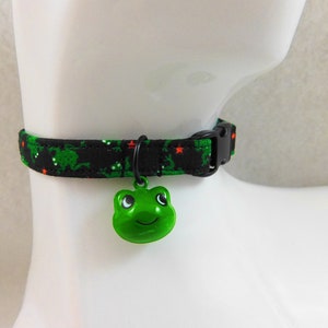 May include: A black cat collar with a green frog pattern and a green frog-shaped bell. The collar has a black plastic buckle and is displayed on a white mannequin head.