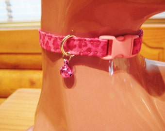 May include: A pink cat collar with a repeating leopard print pattern. The collar has a pink plastic buckle and a gold-colored metal ring holding a small, pink bell. The collar is displayed on a mannequin.