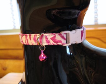May include: A pink and white zebra print cat collar with a white plastic buckle and a small, pink bell. The collar is on a black surface.