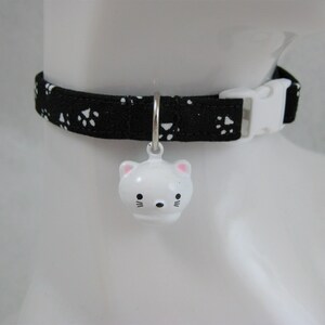K&ouml;nnte beinhalten: Ein schwarzes Katzenhalsband mit wei&szlig;en Pfotenabdr&uuml;cken und einer wei&szlig;en Kunststoffschnalle. Eine wei&szlig;e, katzenf&ouml;rmige Glocke mit rosa Akzenten h&auml;ngt am Halsband. Das Halsband ist f&uuml;r ein Haustier bestimmt.