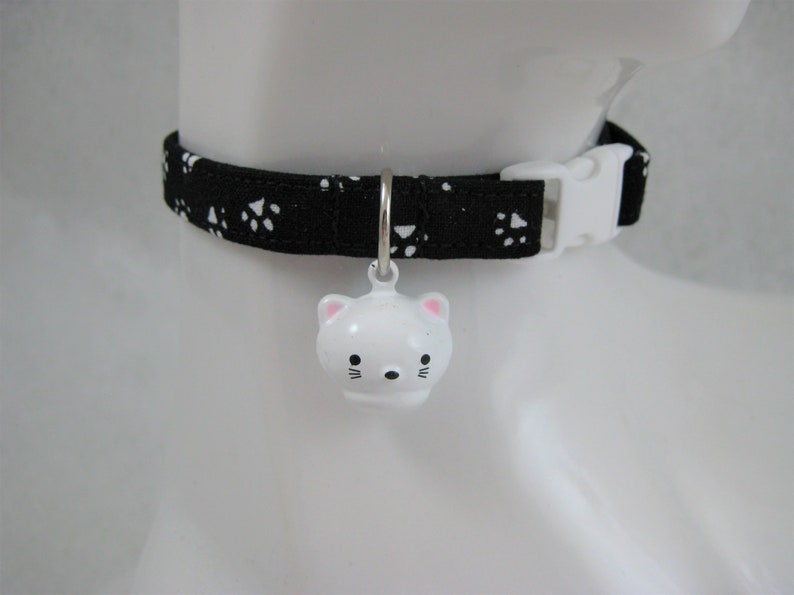 K&ouml;nnte beinhalten: Ein schwarzes Katzenhalsband mit wei&szlig;en Pfotenabdr&uuml;cken und einer wei&szlig;en Kunststoffschnalle. Eine wei&szlig;e, katzenf&ouml;rmige Glocke mit rosa Akzenten h&auml;ngt am Halsband. Das Halsband ist f&uuml;r ein Haustier bestimmt.