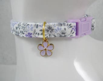 May include: A floral patterned cat collar with a lavender flower charm. The collar features a white background with purple and gray floral designs. It has a lavender plastic buckle and a gold-colored charm holder.