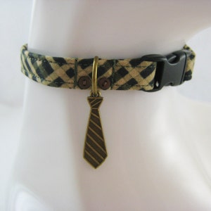 May include: A patterned dog collar with a black plastic buckle. The collar is tan and green with a plaid design. A bronze-colored tie charm hangs from the collar.